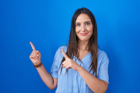 Photo for Young brunette woman standing over blue background smiling and looking at the camera pointing with two hands and fingers to the side. - Royalty Free Image