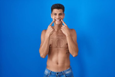 Photo for Young hispanic man standing shirtless over blue background smiling with open mouth, fingers pointing and forcing cheerful smile - Royalty Free Image