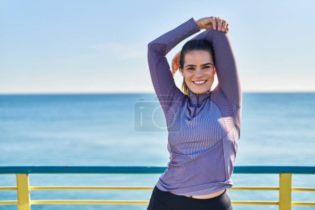 Téléchargez les photos : Jeune belle femme hispanique portant des vêtements de sport étirant le bras au bord de la mer - en image libre de droit