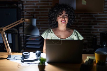 Téléchargez les photos : Jeune femme brune aux cheveux bouclés travaillant au bureau la nuit clin d'oeil en regardant la caméra avec expression sexy, visage joyeux et heureux. - en image libre de droit
