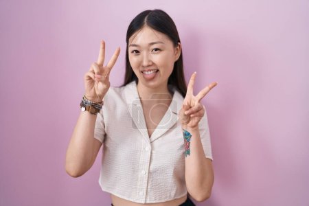 Foto de Mujer joven china de pie sobre fondo rosa sonriendo con la lengua hacia fuera mostrando los dedos de ambas manos haciendo signo de victoria. número dos. - Imagen libre de derechos