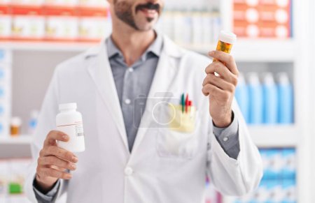 Téléchargez les photos : Young hispanic man pharmacist holding pills bottles at pharmacy - en image libre de droit