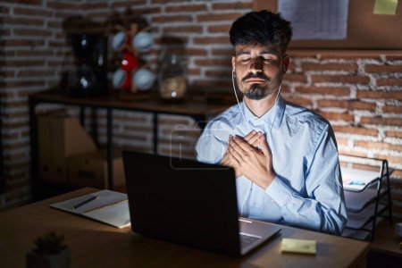 Photo for Young hispanic man with beard working at the office at night smiling with hands on chest with closed eyes and grateful gesture on face. health concept. - Royalty Free Image