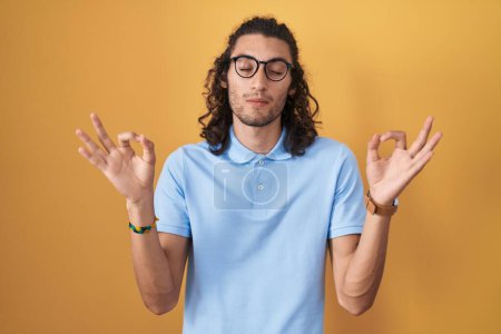 Photo for Young hispanic man standing over yellow background relaxed and smiling with eyes closed doing meditation gesture with fingers. yoga concept. - Royalty Free Image