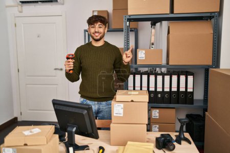 Foto de Hombre árabe con barba trabajando en el comercio de la pequeña empresa sosteniendo el escáner de código de barras sonriendo feliz señalando con la mano y el dedo a un lado - Imagen libre de derechos
