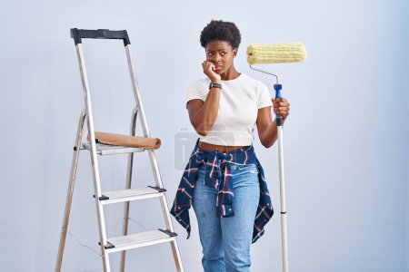 Foto de Mujer afroamericana sosteniendo rodillo pintor buscando estresado y nervioso con las manos en la boca mordiendo las uñas. problema de ansiedad. - Imagen libre de derechos