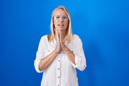 Foto de Young caucasian woman standing over blue background begging and praying with hands together with hope expression on face very emotional and worried. begging. - Imagen libre de derechos