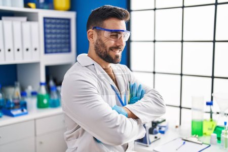 Photo for Young hispanic man wearing scientist uniform sitting with arms crossed gesture at laboratory - Royalty Free Image