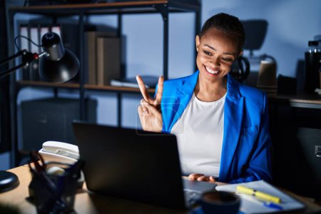 Téléchargez les photos : Belle femme afro-américaine travaillant au bureau la nuit en souriant à la caméra montrant des doigts faisant signe de victoire. numéro deux. - en image libre de droit