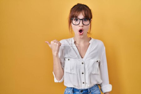 Photo for Young beautiful woman wearing casual shirt over yellow background surprised pointing with hand finger to the side, open mouth amazed expression. - Royalty Free Image