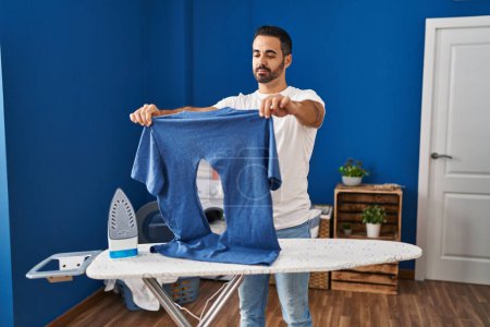 Photo for Young hispanic man with beard ironing holding burned iron shirt at laundry room relaxed with serious expression on face. simple and natural looking at the camera. - Royalty Free Image