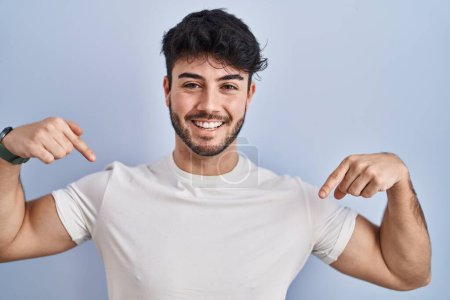 Foto de Hombre hispano con barba de pie sobre fondo blanco mirando confiado con sonrisa en la cara, señalándose con los dedos orgullosos y felices. - Imagen libre de derechos
