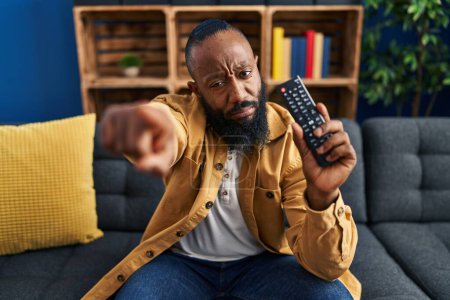 Foto de Hombre afroamericano sosteniendo el control remoto de la televisión señalando con el dedo a la cámara y a usted, gesto de confianza mirando en serio - Imagen libre de derechos