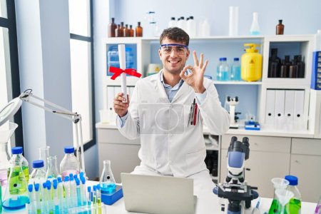 Téléchargez les photos : Jeune homme hispanique travaillant au laboratoire scientifique titulaire d'un diplôme faire ok signe avec les doigts, sourire geste amical excellent symbole - en image libre de droit