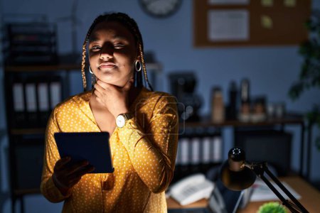 Foto de Mujer afroamericana con trenzas que trabajan en la oficina por la noche con tabletas tocando el cuello doloroso, dolor de garganta para la gripe, la clod y la infección - Imagen libre de derechos
