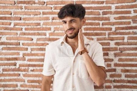 Foto de Hombre árabe con barba de pie sobre ladrillos fondo de la pared tocando boca con mano con expresión dolorosa debido a dolor de muelas o enfermedad dental en los dientes. dentista - Imagen libre de derechos