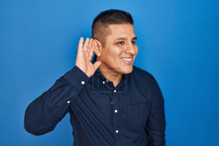 Téléchargez les photos : Jeune homme hispanique debout sur fond bleu souriant avec la main sur l'oreille en écoutant une rumeur ou des commérages. concept de surdité. - en image libre de droit