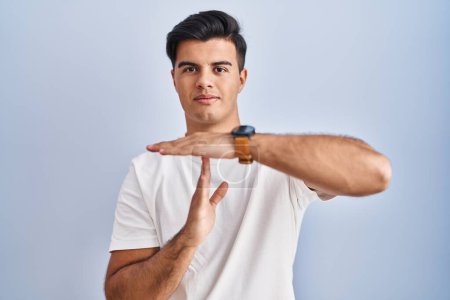 Foto de Hombre hispano de pie sobre fondo azul haciendo un gesto de tiempo fuera con las manos, rostro frustrado y serio - Imagen libre de derechos