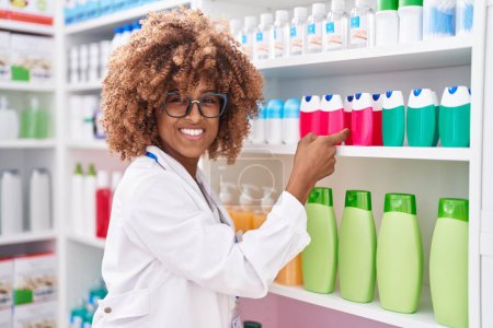 Foto de Farmacéutica afroamericana sonriendo confiada sosteniendo el producto en estanterías en la farmacia - Imagen libre de derechos