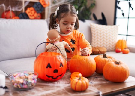 Photo for Adorable hispanic girl wearing pumpkin costume holding baby doll at home - Royalty Free Image