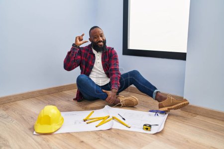 Téléchargez les photos : Homme afro-américain assis sur le sol à la nouvelle maison en regardant les plans de geste souriant et confiant avec la main faisant signe de petite taille avec les doigts regardant et la caméra. concept de mesure. - en image libre de droit