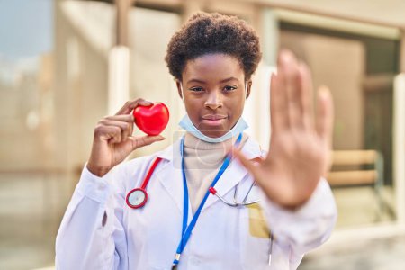 Téléchargez les photos : Femme afro-américaine portant l'uniforme de médecin tenant le cœur avec la main ouverte faisant stop signe avec expression sérieuse et confiante, geste de défense - en image libre de droit