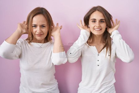 Photo for Middle age mother and young daughter standing over pink background trying to hear both hands on ear gesture, curious for gossip. hearing problem, deaf - Royalty Free Image