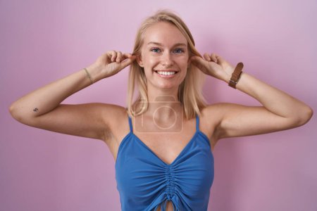 Téléchargez les photos : Jeune femme caucasienne debout sur fond rose souriant tirant les oreilles avec les doigts, geste drôle. problème d'audition - en image libre de droit