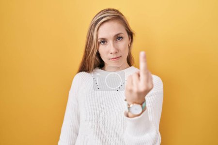 Foto de Young caucasian woman wearing white sweater over yellow background showing middle finger, impolite and rude fuck off expression - Imagen libre de derechos