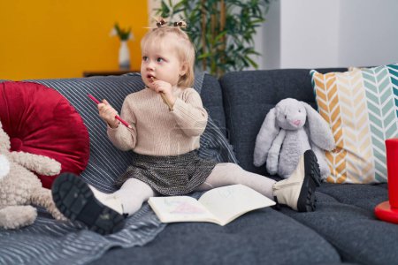 Photo for Adorable blonde girl preschool student sitting on sofa drawing on notebook at home - Royalty Free Image