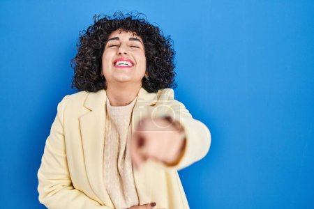 Photo for Young brunette woman with curly hair standing over blue background laughing at you, pointing finger to the camera with hand over body, shame expression - Royalty Free Image