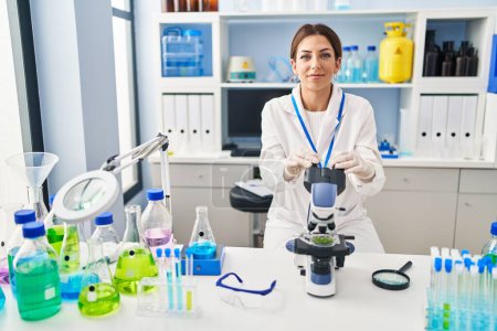 Foto de Mujer hispana joven vistiendo uniforme científico usando microscopio en laboratorio - Imagen libre de derechos