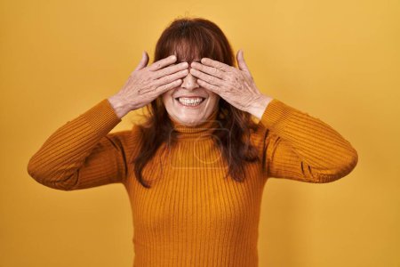 Téléchargez les photos : Femme hispanique du Moyen Âge debout sur fond jaune couvrant les yeux avec les mains souriantes gaies et drôles. concept aveugle. - en image libre de droit