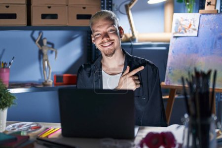 Foto de Joven hombre caucásico usando portátil por la noche en el estudio de arte alegre con una sonrisa de la cara señalando con la mano y el dedo hacia el lado con expresión feliz y natural en la cara - Imagen libre de derechos