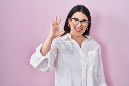 Photo for Young brunette woman standing over pink background smiling positive doing ok sign with hand and fingers. successful expression. - Royalty Free Image