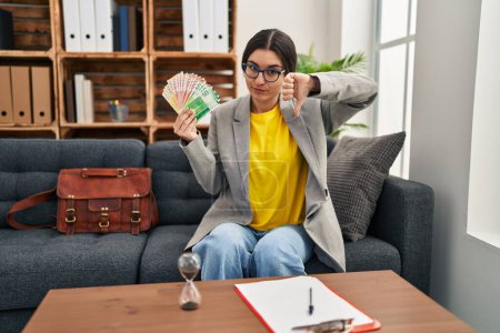 Photo for Young hispanic woman working at consultation office holding money with angry face, negative sign showing dislike with thumbs down, rejection concept - Royalty Free Image