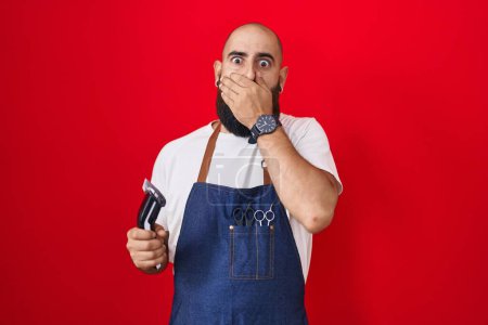 Photo for Young hispanic man with beard and tattoos wearing barber apron holding razor shocked covering mouth with hands for mistake. secret concept. - Royalty Free Image