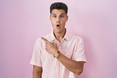 Foto de Joven hombre hispano de pie sobre fondo rosa sorprendido señalando con el dedo hacia un lado, boca abierta expresión asombrada. - Imagen libre de derechos