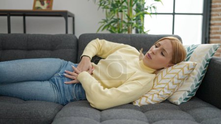 Téléchargez les photos : Young blonde woman suffering for menstrual pain lying on sofa at home - en image libre de droit
