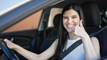 Téléchargez les photos : Jeune belle hispanique femme assise sur la voiture faisant pouce vers le haut geste à la rue - en image libre de droit