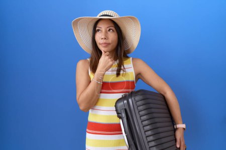 Foto de Mujer china de mediana edad sosteniendo maleta va en vacaciones de verano cara seria pensando en la pregunta con la mano en la barbilla, pensativo acerca de la idea confusa - Imagen libre de derechos