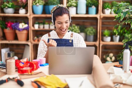 Téléchargez les photos : Young hispanic woman working at florist shop doing video call pointing thumb up to the side smiling happy with open mouth - en image libre de droit
