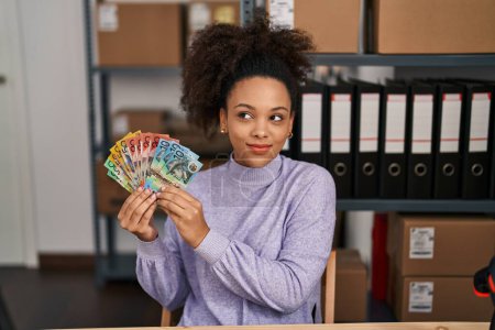Téléchargez les photos : Jeune femme afro-américaine travaillant dans une petite entreprise e-commerce tenant des dollars australiens souriant regardant vers le côté et regardant loin en pensant. - en image libre de droit