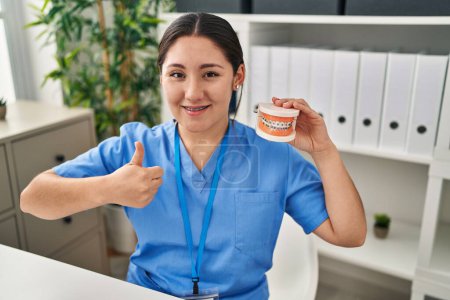 Photo for Young latin dentist woman holding denture with braces smiling happy and positive, thumb up doing excellent and approval sign - Royalty Free Image