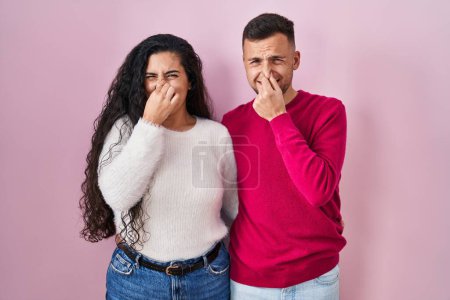 Téléchargez les photos : Jeune couple hispanique debout sur fond rose sentant quelque chose de puant et dégoûtant, odeur intolérable, retenant souffle avec les doigts sur le nez. mauvaise odeur - en image libre de droit