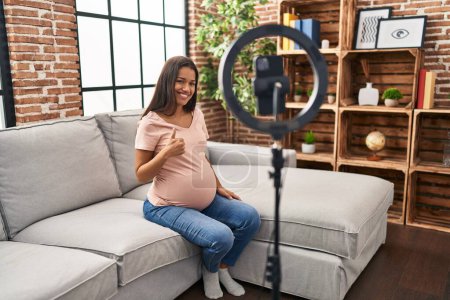Téléchargez les photos : Jeune femme enceinte enregistrement tutoriel vlog avec smartphone à la maison souriant heureux et positif, pouce levé faire excellent et signe d'approbation - en image libre de droit