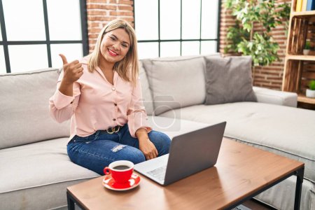 Photo for Young hispanic woman using laptop sitting on the sofa at home doing happy thumbs up gesture with hand. approving expression looking at the camera showing success. - Royalty Free Image