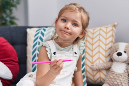 Photo for Adorable blonde girl preschool student sitting on sofa holding color pencil at home - Royalty Free Image