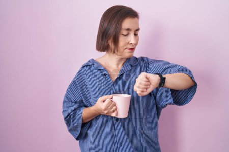 Foto de Mujer hispana de mediana edad bebiendo una taza de café comprobando la hora en el reloj de muñeca, relajada y confiada - Imagen libre de derechos