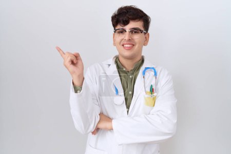 Photo for Young non binary man wearing doctor uniform and stethoscope with a big smile on face, pointing with hand and finger to the side looking at the camera. - Royalty Free Image